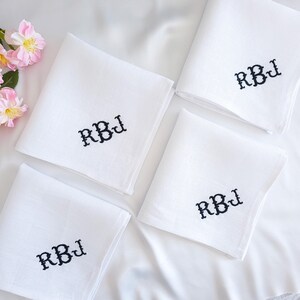 May include: Four square white fabric handkerchiefs, each featuring the embroidered black monogram letters RBJ in a decorative font. The items are arranged in a group, with a cluster of pink and yellow flowers visible in the upper left corner.