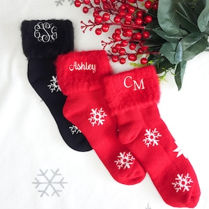 May include: One black and two red knit stockings featuring white snowflake patterns and fuzzy cuffs. The black stocking is embroidered with the monogram ESG, while the red stockings are personalized with the names Ashley and CM in white script.