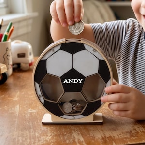 以下が含まれることがあります： サッカーボールの形をした円形の木製貯金箱で、前面は透明なパネルになっています。黒と白の六角形の模様が特徴で、中央には白い文字でANDYと印刷されています。上部のスロットから硬貨を中に入れることができます。