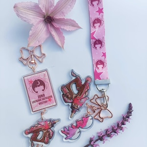 Op de afbeelding: Een roze sleutelhanger-set met een stoffen polsband met ster- en karakter-motieven, een rechthoekige acryl bedel met de tekst JUNO LOCKING SATELLITE VECTOR, twee pistoolvormige bedels en twee haai-vormige bedels, verbonden door roségouden strikjes.