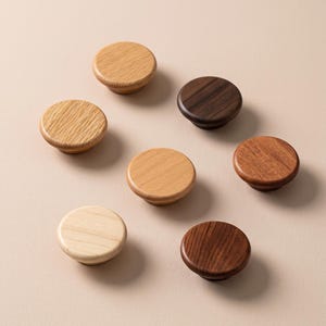 Könnte beinhalten: Eine Ansammlung von sieben runden Schrankknöpfen aus Holz, die in einer Gruppe angeordnet sind. Jeder Knopf weist eine eigene Holzmaserung und Farbe auf, die von hellem Beige und blassem Eichenholz bis zu mittleren Brauntönen und dunklem Walnuss reichen.