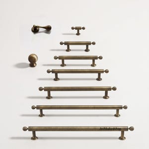 Op de afbeelding: Een collectie kastbeslag van antiek messing, inclusief een ronde knop, een hefboomgreep en zes staafgrepen van verschillende lengtes. Elk stuk is voorzien van een geribbeld, bolvormig einddetail. De tekst "Joshbakedshop" is zichtbaar op de onderste greep.