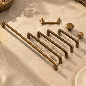 May include: A collection of antique brass cabinet hardware including one long pull handle, four medium-length pull handles, one small pull handle, and two round knobs. Each piece features a smooth, matte finish with a warm, metallic golden-brown tone.