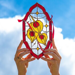 Puede incluir: Un atrapasoles de vitral ovalado con un borde rojo y un diseño central de tres vainas de semillas amarillas con centros rojos. Las piezas de vidrio están unidas por líneas de soldadura de metal oscuro y un patrón de rejilla de diamantes.
