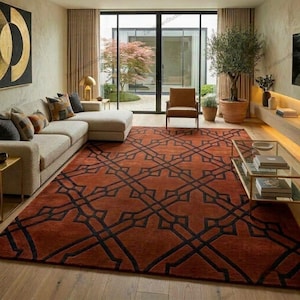May include: A large rectangular area rug featuring a deep rust-orange background with a bold, black geometric lattice pattern. The rug covers the floor of a living room, extending from a light-colored sectional sofa to a glass-topped coffee table.