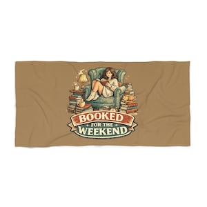 Könnte beinhalten: Ein beigefarbenes Handtuch mit einer grafischen Illustration einer Person, die in einem blaugrünen Sessel liest, umgeben von Bücherstapeln, einer Lampe, einer Tasse und einer Katze. Der Text "BOOKED FOR THE WEEKEND" steht auf einem rot-blauen Banner.