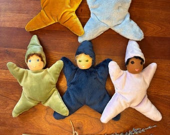 May include: Five star-shaped plush dolls with pointed hats, each featuring a different color: mustard yellow, light blue, sage green, navy blue, and pale lavender. Each doll has a soft fabric body and a simple embroidered face. A bundle of dried lavender stems tied with twine is positioned below the dolls.