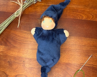 May include: A soft plush doll wearing a dark blue hooded sleeper with a knotted bottom. The doll has light skin, brown hair, and embroidered facial features. Beside the doll are dried lavender sprigs and a sewing needle with green thread.