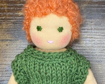 May include: A handmade fabric doll with curly orange hair, a light beige complexion, and pink cheeks. The doll features embroidered green eyes and a small pink mouth. It is wearing a short-sleeved, olive green knitted sweater with a ribbed texture.