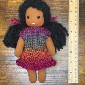 May include: A handmade fabric doll with dark skin, black yarn hair tied with magenta ribbons, and a knitted dress featuring a gradient of purple, blue, and orange. A wooden ruler placed beside the doll shows a total height of approximately 7.5 inches.