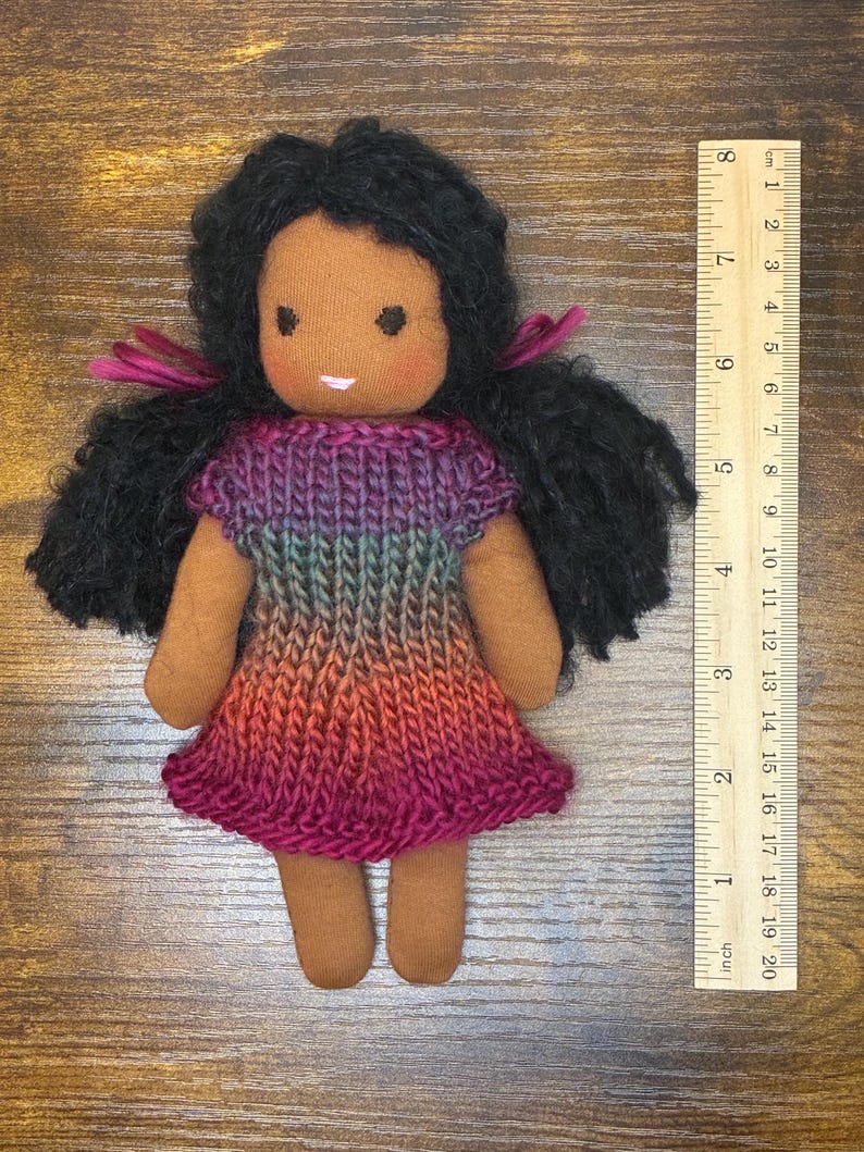 May include: A handmade fabric doll with dark skin, black yarn hair tied with magenta ribbons, and a knitted dress featuring a gradient of purple, blue, and orange. A wooden ruler placed beside the doll shows a total height of approximately 7.5 inches.