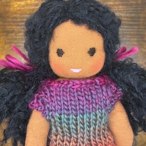 May include: A handmade fabric doll with dark skin and curly black yarn hair tied with magenta ribbons. The doll features embroidered dark eyes, rosy cheeks, and a small pink mouth. It wears a knitted dress with a gradient of purple, blue, and pink.