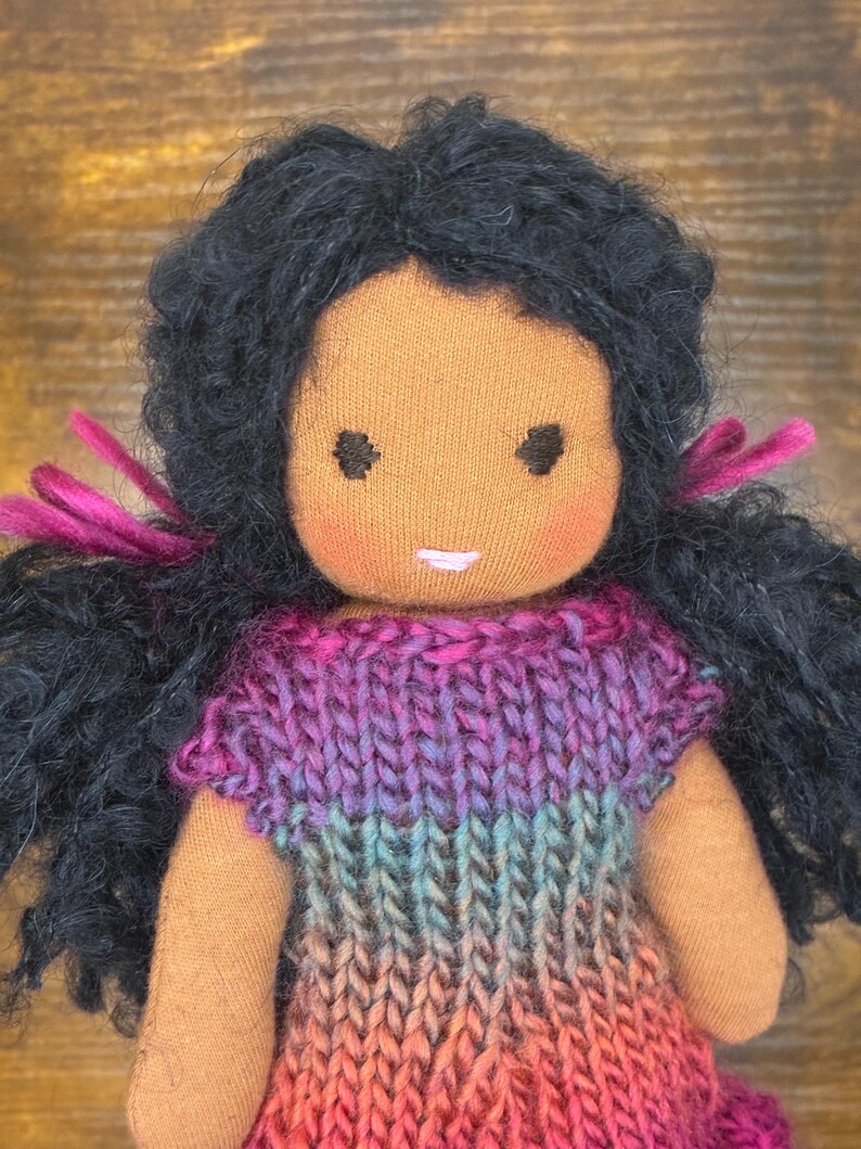 May include: A handmade fabric doll with dark skin and curly black yarn hair tied with magenta ribbons. The doll features embroidered dark eyes, rosy cheeks, and a small pink mouth. It wears a knitted dress with a gradient of purple, blue, and pink.
