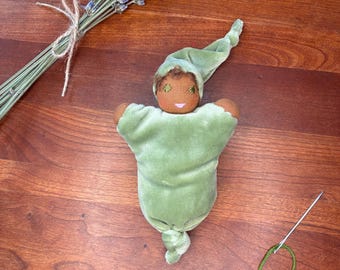 May include: A soft fabric doll with dark skin, wearing a light green velvet sleeper with a matching pointed cap. The doll has embroidered facial features and a knotted bottom. Beside the doll are dried lavender sprigs and a sewing needle with green thread.