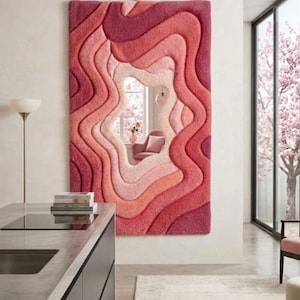 May include: A rectangular wall-mounted mirror framed by a thick, textured, tufted textile border featuring concentric, wavy layers in various shades of pink, ranging from deep magenta to soft blush, suspended from a dark horizontal metal rod.
