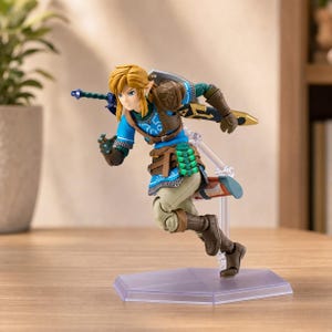 Op de afbeelding: Een beweegbaar actiefiguur van een blond personage in een blauwe tuniek met witte patronen, bruin harnas en een beige broek. Het figuur staat op een doorzichtige zeshoekige standaard en houdt een zwaard en een schild vast.