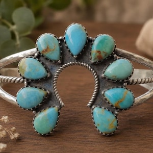 May include: A silver cuff bracelet featuring a horseshoe-shaped centerpiece adorned with nine teardrop-shaped turquoise stones. The metal band has a hammered texture and a twisted rope detail surrounding the stones, which display blue and green hues.
