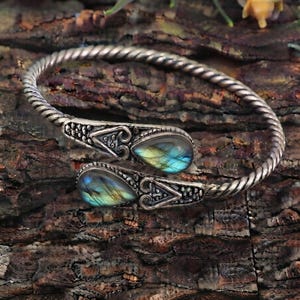 Può includere: Un bracciale a fascia in metallo color argento con un design a banda intrecciata. Le due estremità aperte sono decorate con pietre preziose di labradorite a forma di goccia che mostrano riflessi blu e oro, incastonate in montature in metallo lavorato.