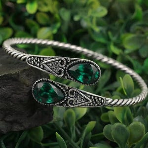 May include: A silver-toned open cuff bracelet featuring a twisted metal band and two teardrop-shaped dark green gemstones. Each gemstone is set within an ornate, textured metal frame that tapers to a point, creating a symmetrical design.