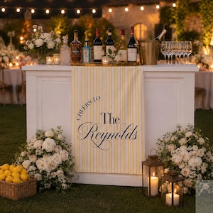 May include: A white bar counter featuring a yellow and white striped fabric banner with the text Cheers to The Reynolds in dark blue script. The bar is topped with wine bottles, a metal ice bucket, and glasses, flanked by white floral arrangements.