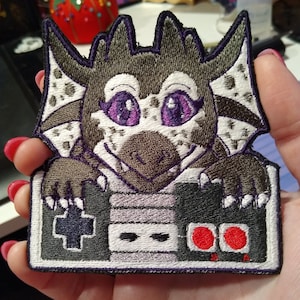 Puede incluir: Parche bordado con un dragón de dibujos animados con un mando. El dragón tiene ojos morados y es marrón, gris y blanco con detalles morados. El mando es gris con una cruz negra, un rectángulo blanco y botones rojos.