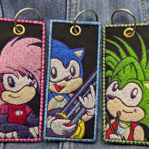 May include: Three embroidered keychains featuring cartoon hedgehog characters. Each keychain has a black background with a different character in vibrant colours. The keychains have a metal ring for attachment.