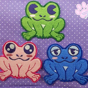 May include: Three embroidered frog patches in green, pink, and blue. Each frog has large, expressive eyes and a smiling or thoughtful expression. The patches are on a purple polka dot background, showcasing a cute, whimsical design.