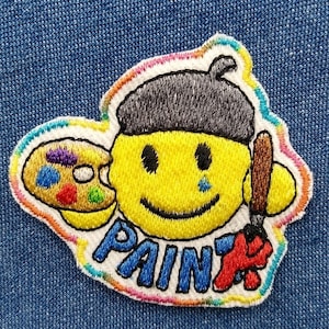 May include: Embroidered patch featuring a smiling yellow face wearing a gray beret and holding a paintbrush and a paint palette. The word "PAINT" is embroidered in blue and red. The patch has a rainbow border and is set against a blue denim background.