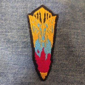 Op de afbeelding: Geborduurde patch in schildvorm, met een ontwerp in geel, blauw en rood. De patch heeft een zwarte rand en is geplaatst tegen een denim achtergrond. Het ontwerp omvat verticale lijnen en een vlamachtig element.