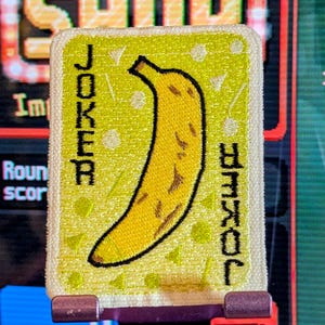 Puede incluir: Un parche rectangular con una ilustración de plátano amarillo sobre un fondo verde claro. La palabra "JOKER" está impresa verticalmente a ambos lados del plátano. El parche tiene un borde blanco y está montado sobre una superficie morada y roja.