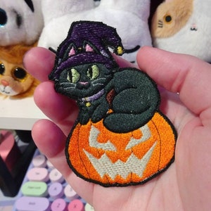 Może przedstawiać: Haftowana naszywka przedstawiająca czarnego kota w fioletowym kapeluszu czarownicy, siedzącego na pomarańczowej dyni. Kot ma zielone oczy i fioletowy kołnierz. Rozmyte tło z pluszowymi zabawkami.