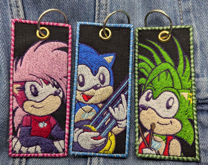 May include: Three embroidered keychains featuring cartoon hedgehog characters. Each keychain has a black background with a different character in vibrant colours. The keychains have a metal ring for attachment.