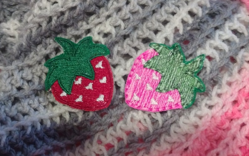 Op de afbeelding: Twee geborduurde aardbeienpatches. E&eacute;n is rood met witte zaadjes en een groen blad, de andere is roze met witte zaadjes en een groen blad. De patches liggen op een gehaakte achtergrond in grijs, wit en roze.