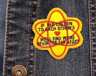 May include: A yellow and red embroidered patch with the text "IF WE LISTEN TO EACH OTHER'S WE'LL FIND WE'RE NEVER TOO FAR APART" is sewn onto a denim jacket. The jacket has two metal buttons.