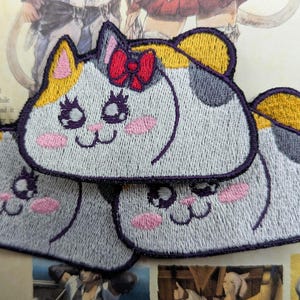 May include: Embroidered patches featuring a cute cat design. The patches are gray, white, and yellow with pink cheeks, purple eyes, and a red bow. The patches are stacked on top of each other.