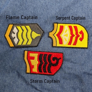 Peut inclure: Trois écussons brodés avec les textes "Flame Captain", "Serpent Captain" et "Storm Captain". Les écussons présentent des motifs jaunes, rouges, noirs et gris sur un fond en denim bleu. Les écussons sont de formes variées.