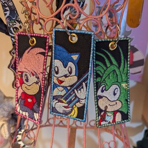 May include: Three embroidered keychains featuring Sonic the Hedgehog characters. Each keychain has a black background with a character design, a gold-coloured ring, and a coloured border. The characters include Amy Rose, Sonic, and another character.