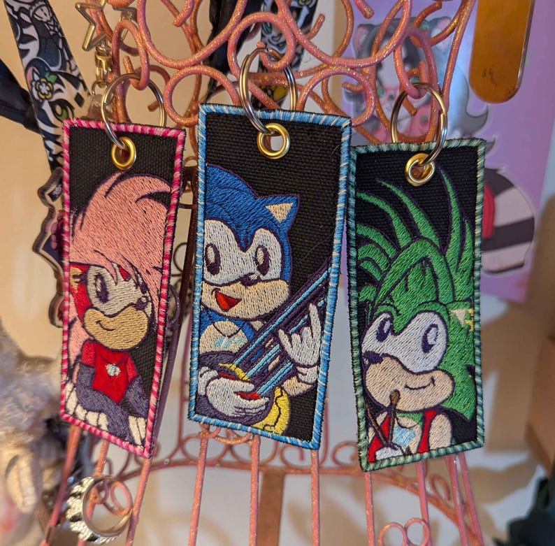 May include: Three embroidered keychains featuring Sonic the Hedgehog characters. Each keychain has a black background with a character design, a gold-coloured ring, and a coloured border. The characters include Amy Rose, Sonic, and another character.