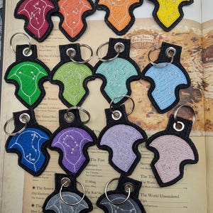 K&ouml;nnte beinhalten: Eine Sammlung von vierzehn schildf&ouml;rmigen, bestickten Schl&uuml;sselanh&auml;ngern in verschiedenen Farben wie Rot, Orange, Gelb, Gr&uuml;n, Blau, Lila und Grau. Jeder Schl&uuml;sselanh&auml;nger zeigt ein wei&szlig;es Sternbildmuster und einen silbernen Metallring.