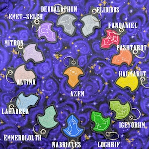 K&ouml;nnte beinhalten: Eine Sammlung von dreizehn bestickten Schl&uuml;sselanh&auml;ngern in verschiedenen Farben, jeder mit einem einzigartigen Sternbild-Design und einem Metallring. Die Anh&auml;nger sind mit Namen wie Azem, Emet-Selch, Deudalaphon, Elidibus, Fandaniel, Pashtarot, Halmarut, Lahabrea, Emmeroloth, Nabriales, Igeyorhm, Mitron und Loghrif beschriftet.