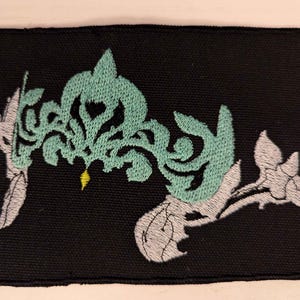 May include: Black rectangular patch with rounded corners, featuring an embroidered design. The design includes a teal crown-like shape, white floral elements, and a small yellow accent. The patch is likely for clothing or accessories.