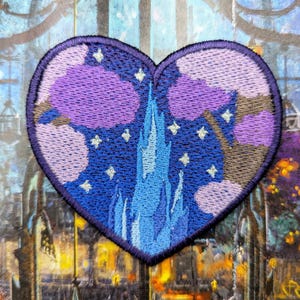 May include: Embroidered heart-shaped patch with a castle scene, purple trees, and stars on a dark blue backdrop. The patch has a dark blue border, ideal for customising clothing or accessories.