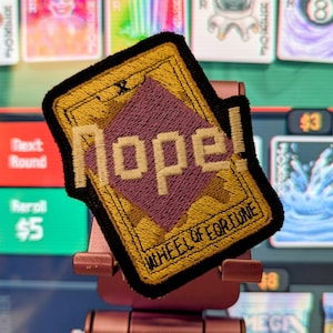 Può includere: Toppa ricamata con la parola "Nope!" in lettere pixelate, su una forma a diamante viola e uno sfondo dorato. La toppa ha un bordo nero e le parole "WHEEL OF FORTUNE" in basso.