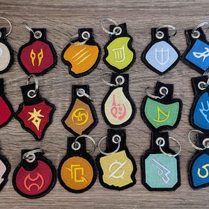 Op de afbeelding: Een verzameling geborduurde sleutelhangers met diverse kleurrijke symbolen en ontwerpen. Elke sleutelhanger heeft een zwarte achterkant, een zilveren sleutelring en een uniek embleem in tinten rood, geel, blauw, groen en wit. De sleutelhangers zijn gerangschikt op een houten oppervlak.
