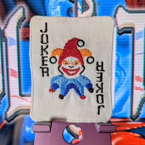 May include: Embroidered playing card featuring a smiling joker with a red hat, blue outfit, and orange accents. The word "JOKER" is embroidered on either side of the image. The card is displayed on a purple stand.