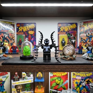 Puede incluir: Una minifigura de Venom en blanco y negro con cuatro tentáculos negros articulados en la espalda, situada entre un pequeño cristal verde translúcido bajo una cúpula de cristal y un reloj de bolsillo de estilo antiguo con el texto SYMBIOST.