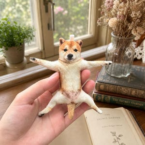 Puede incluir: Un pequeño adorno de perro Shiba Inu de fieltro con pelaje beige y blanco, nariz negra y ojos oscuros. La figura se muestra en una pose extendida con las cuatro extremidades hacia afuera y una pequeña cola, con un lazo de tela blanca en la parte superior.