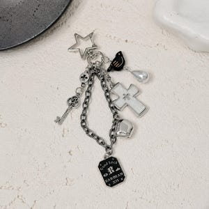 Puede incluir: Un llavero de metal plateado con un cierre en forma de estrella, un dije de pájaro negro, una cruz blanca, una llave plateada, un corazón y una etiqueta rectangular negra con el texto Good Luck BARDIAN XII impreso en blanco.