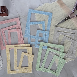 Könnte beinhalten: Eine Sammlung rechteckiger Papierrahmen in Pastellrosa, Blau, Gelb, Grün und Grau, jeweils mit einer strukturierten Kreuzschraffur. Drei dunkelbraune Holzblöcke sind mit den Buchstaben A, E und M markiert, ergänzt durch eine Schere.