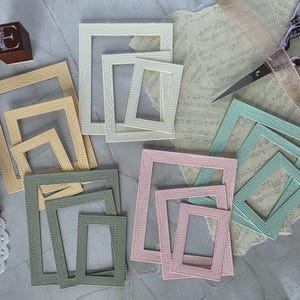 Könnte beinhalten: Eine Sammlung rechteckiger Papierrahmen in verschiedenen Pastellfarben wie Creme, Gelb, Salbeigrün und Hellrosa. Jeder Rahmen weist ein strukturiertes, gepunktetes Randmuster auf. Die Rahmen sind in ineinander verschachtelten Dreiersets angeordnet.